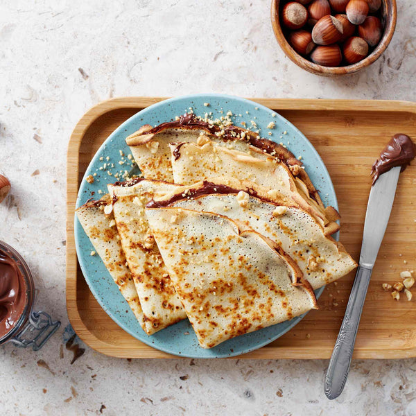 Crepes on a blue plate filled with Nutella chocolate spread and hazelnuts on a tray with a knife covered in chocolate and next to a bowl of hazelnuts